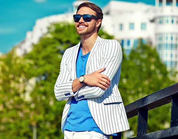 Hombre guapo inconformista sonriente divertido en elegante traje blanco de verano posando en la calle con gafas de sol