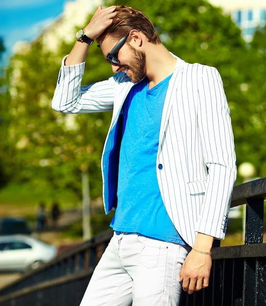 Hombre guapo inconformista sonriente divertido en elegante traje blanco de verano posando en la calle con gafas de sol