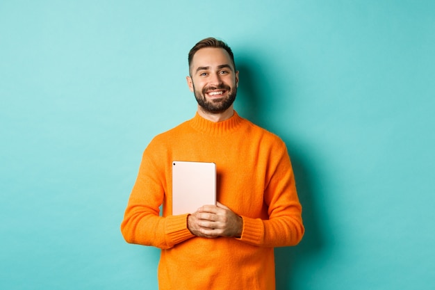 Hombre guapo independiente sosteniendo portátil y sonriendo, feliz de pie sobre la pared turquesa claro