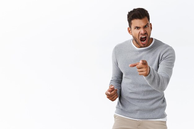 Hombre guapo indignado con mal genio perdiendo el control de las emociones angustiado apuntando a la cámara y gritando acusaciones culpar a alguien con ira y sentimientos furiosos de fondo blanco