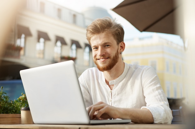 Hombre guapo jengibre adulto con computadora portátil sentado en la terraza de un restaurante o cafetería