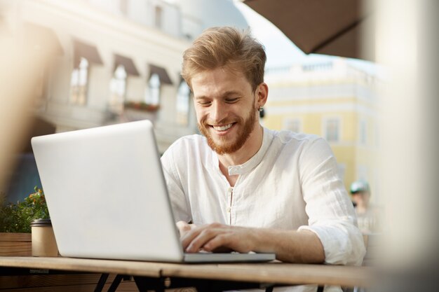 Hombre guapo jengibre adulto con computadora portátil sentado en la terraza de un restaurante o cafetería