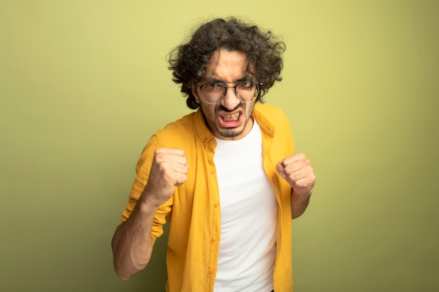 Hombre guapo joven agresivo con gafas mirando al frente apretando los puños aislados en la pared verde oliva