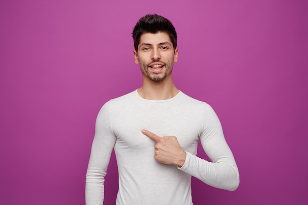 Hombre guapo joven alegre mirando a la cámara apuntándose a sí mismo sobre fondo púrpura