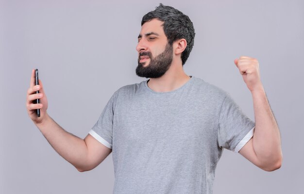 Hombre guapo joven alegre sosteniendo y mirando el teléfono móvil y levantando el puño aislado en blanco