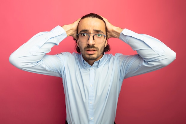 Hombre guapo joven ansioso con gafas manteniendo las manos en la cabeza mirando al frente aislado en la pared rosa