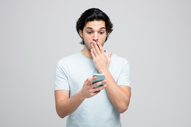 Hombre guapo joven ansioso sosteniendo y mirando el teléfono móvil manteniendo la mano en la boca aislado sobre fondo blanco.