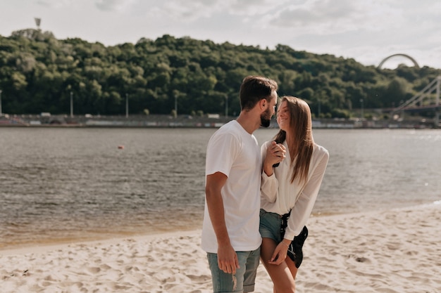 Hombre guapo joven con atractiva mujer de pelo largo caminando en la playa, besos, tomados de la mano