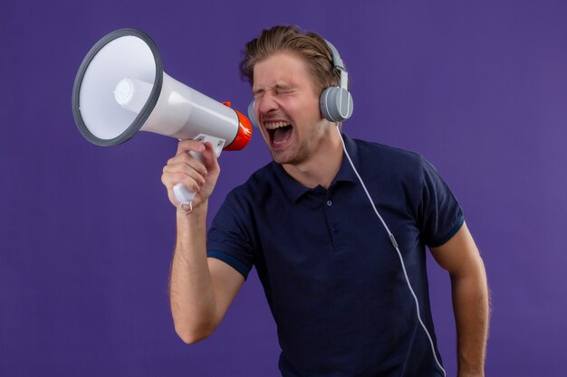 Hombre guapo joven con auriculares gritando por megáfono parado sobre fondo púrpura