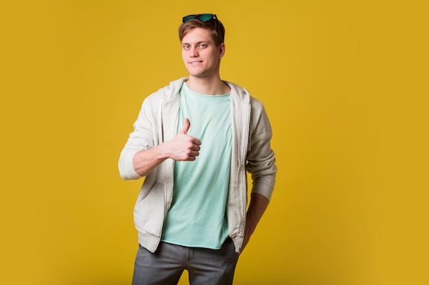 Hombre guapo joven con barba de pie sobre una pared amarilla sonriendo con cara feliz mirando y apuntando hacia el lado con el pulgar hacia arriba.