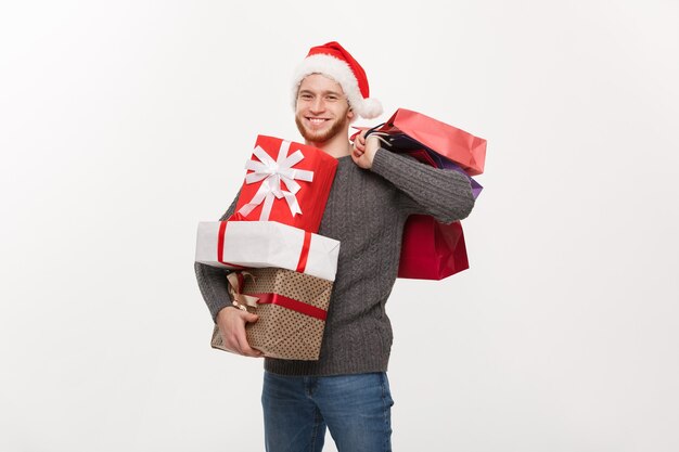 Hombre guapo joven con barba sosteniendo un montón de regalos y bolsas de la compra con expresión facial feliz en blanco