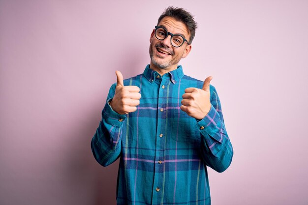 Hombre guapo joven con camisa casual y gafas de pie sobre el signo de éxito de fondo rosa