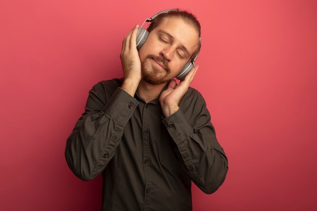 Hombre guapo joven en camisa gris con auriculares en la cabeza disfrutando de su música favorita con los ojos cerrados