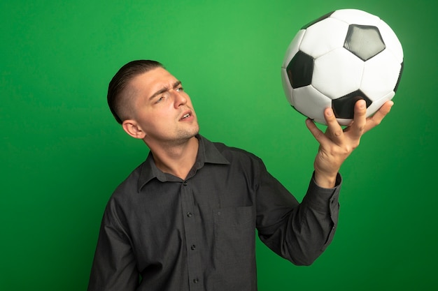 Hombre guapo joven en camisa gris con balón de fútbol mirándolo con cara seria de pie sobre la pared verde