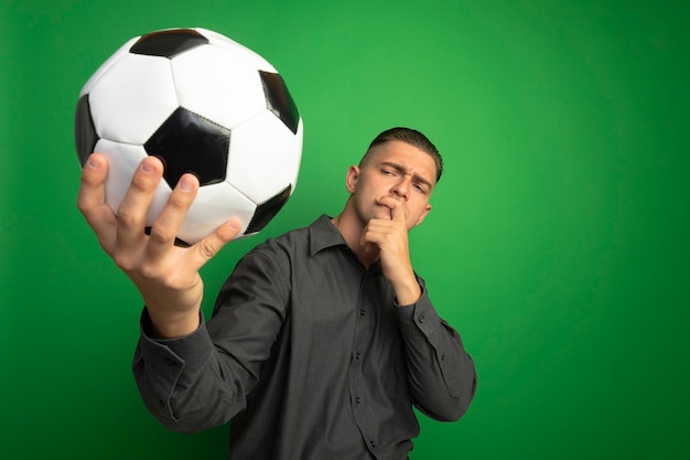 Foto gratuita hombre guapo joven en camisa gris con balón de fútbol mirándolo intrigado