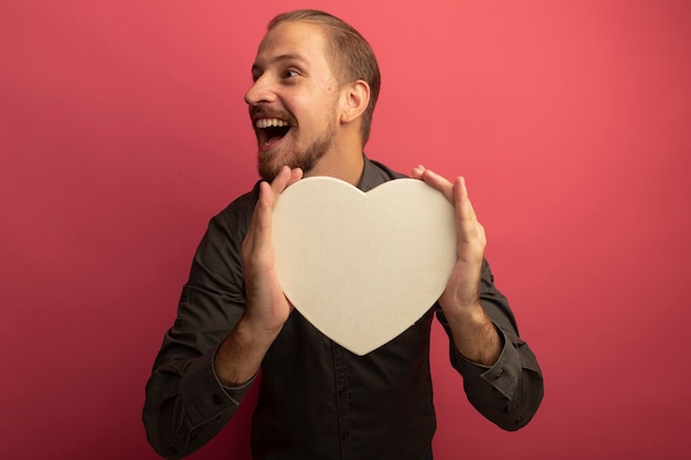 Hombre guapo joven en camisa gris con corazón de cartón mirando a un lado sonriendo alegremente