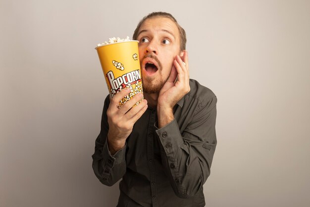 Hombre guapo joven en camisa gris con cubo con palomitas de maíz mirando sorprendido y asombrado