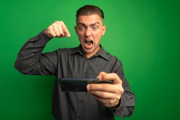 Hombre guapo joven en camisa gris con gadget apretando el puño loco y enojado gritando de pie sobre la pared verde