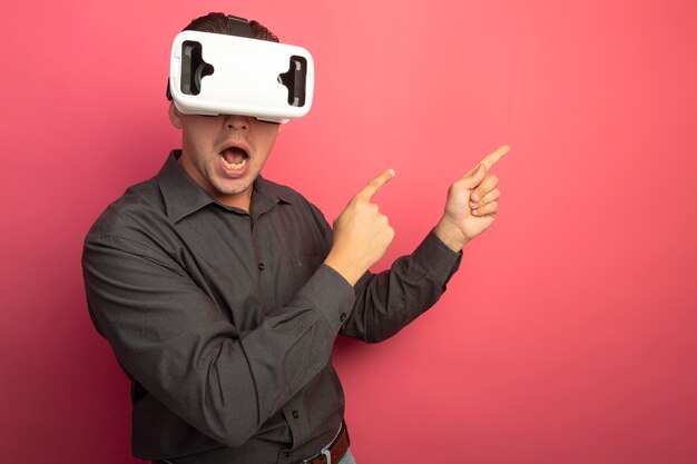 Hombre guapo joven en camisa gris con gafas de realidad virtual mirando al frente apuntando con los dedos hacia el lado parado sobre la pared rosa