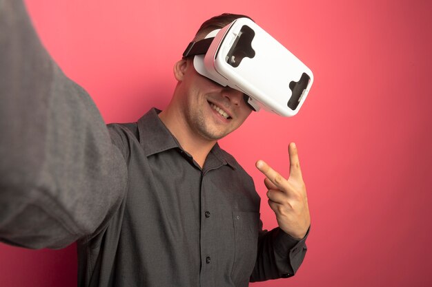 Hombre guapo joven en camisa gris con gafas de realidad virtual sonriendo mostrando signo v parado sobre pared rosa