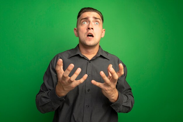 Hombre guapo joven en camisa gris gritando con los brazos hacia fuera loco loco y frustrado de pie sobre la pared verde