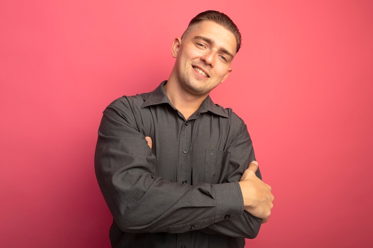 Foto gratuito hombre guapo joven en camisa gris mirando al frente con las manos cruzadas sobre el pecho sonriendo confiado de pie sobre la pared rosa
