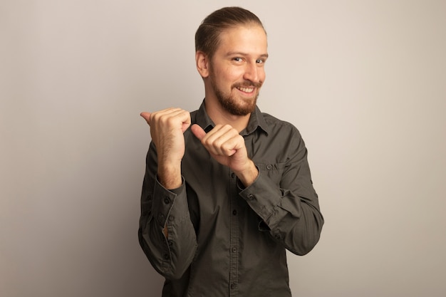 Hombre guapo joven en camisa gris mirando al frente sonriendo señalando confianza con los dedos índices hacia el lado de pie sobre la pared blanca