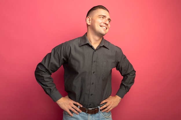 Foto gratuita hombre guapo joven en camisa gris mirando a un lado sonriendo confiado con los brazos en la cadera de pie sobre la pared rosa