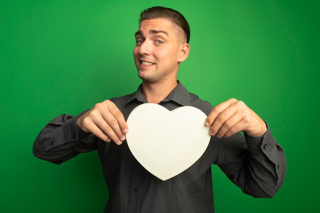 Hombre guapo joven en camisa gris mostrando corazón de cartón mirando al frente sonriendo con cara feliz de pie sobre la pared verde