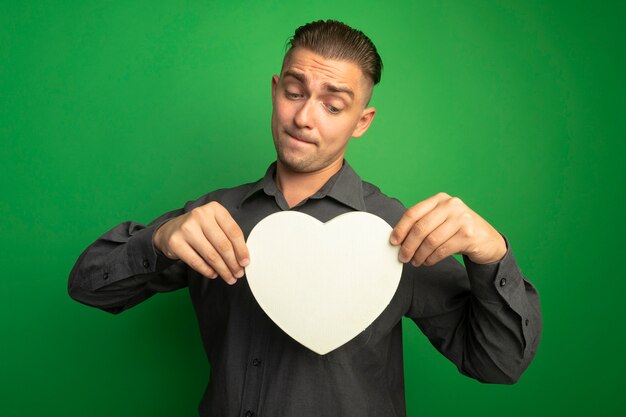 Hombre guapo joven en camisa gris mostrando corazón de cartón mirándolo con expresión confusa de pie sobre la pared verde