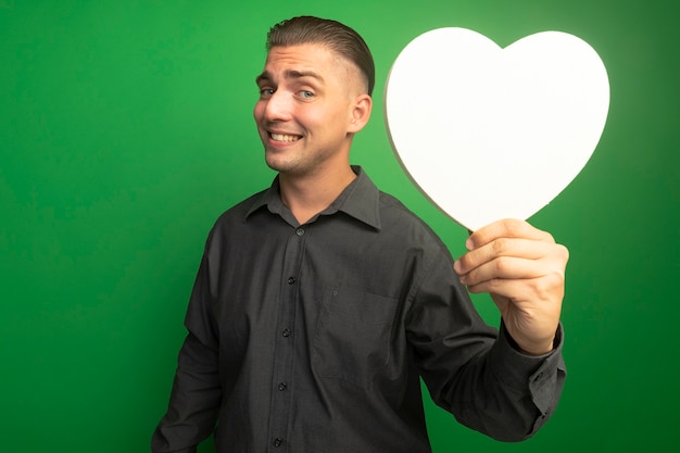 Hombre guapo joven en camisa gris mostrando corazón de cartón sonriendo confiado