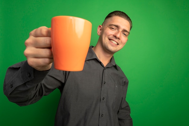 Hombre guapo joven en camisa gris mostrando taza naranja sonriendo con cara feliz de pie sobre la pared verde
