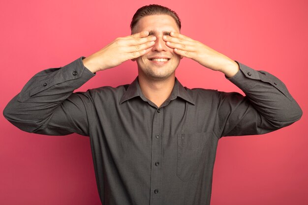 Hombre guapo joven en camisa gris que cubre los ojos con las manos sonriendo alegremente de pie sobre la pared rosa