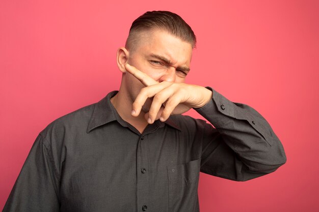 Hombre guapo joven en camisa gris secándose la nariz con la mano de pie sobre la pared rosa