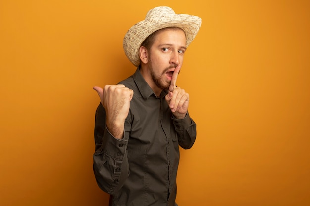 Hombre guapo joven en camisa gris y sombrero de verano apuntando con el pulgar hacia atrás haciendo gesto de silencio con el dedo en los labios