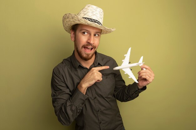 Hombre guapo joven en camisa gris y sombrero de verano con avión de juguete apuntando con el dedo índice feliz y positivo