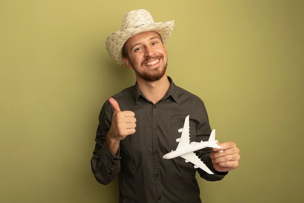 Hombre guapo joven en camisa gris y sombrero de verano con avión de juguete feliz y positivo sonriendo alegremente mostrando los pulgares para arriba