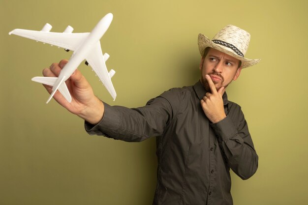 Hombre guapo joven en camisa gris y sombrero de verano con avión de juguete lookign con expresión pensativa en el pensamiento de la cara