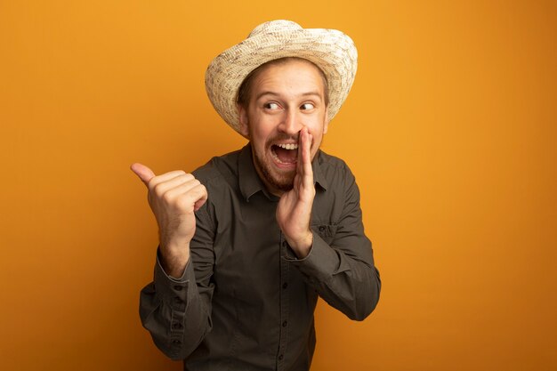 Hombre guapo joven en camisa gris y sombrero de verano contando un secreto con la mano cerca de la boca pointign con el pulgar hacia atrás emocional