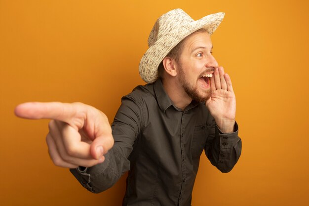 Hombre guapo joven en camisa gris y sombrero de verano gritando o llamando a alguien con la mano cerca de la boca y apuntando con el dedo índice a algo