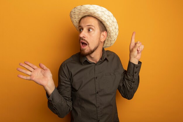 Hombre guapo joven en camisa gris y sombrero de verano mirando a un lado sorprendido y confundido presentando algo con el brazo y el dedo índice de herradura