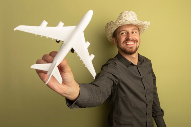 Hombre guapo joven en camisa gris y sombrero de verano mostrando avión de juguete feliz y positivo sonriendo alegremente