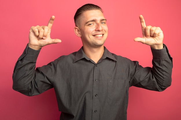 Foto gratuita hombre guapo joven en camisa gris sonriendo alegremente mostrando gesto de tamaño con el símbolo de medida de los dedos de pie sobre la pared rosa