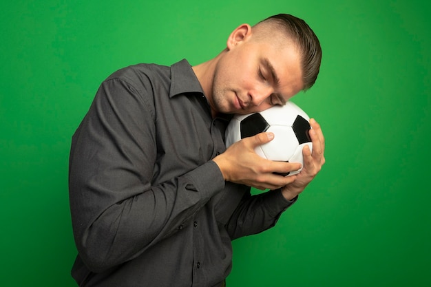 Hombre guapo joven en camisa gris sosteniendo un balón de fútbol con la cabeza apoyada en la bola con los ojos cerrados