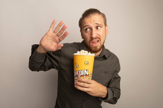 Hombre guapo joven en camisa gris sosteniendo un cubo con palomitas de maíz mirando a un lado preocupado y confundido sosteniendo la mano como gesto de defensa