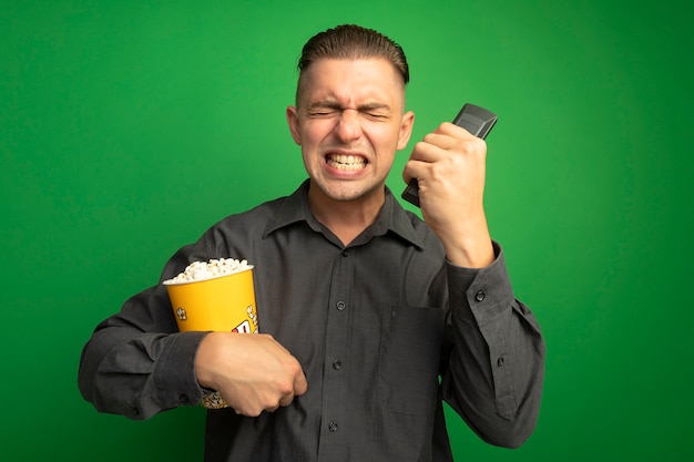 Hombre guapo joven en camisa gris sosteniendo un cubo con palomitas de maíz usando el control remoto de la televisión gritando loco loco y frustrado de pie sobre la pared verde