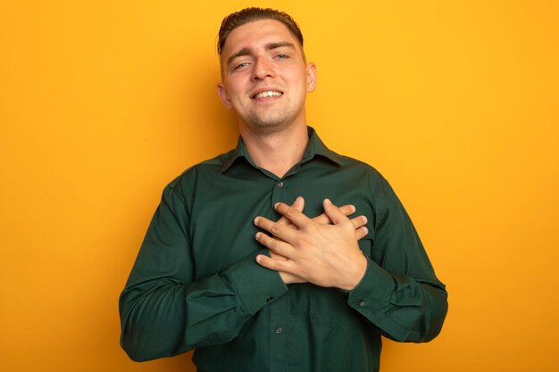 Hombre guapo joven en camisa verde cogidos de la mano en el pecho sintiéndose agradecido