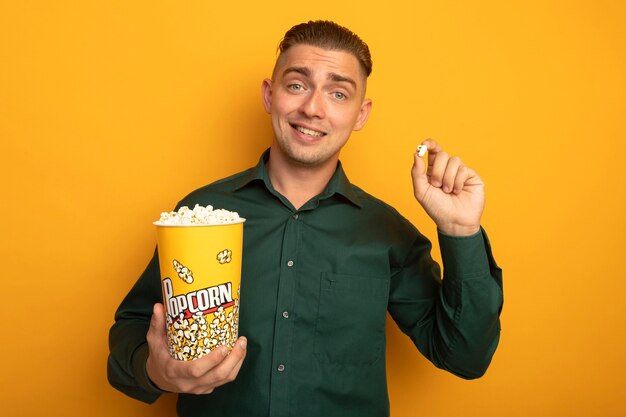 Hombre guapo joven en camisa verde con cubo con palomitas de maíz sonriendo feliz y positivo