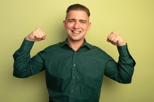 Hombre guapo joven en camisa verde levantando los puños feliz y positivo sonriendo alegremente