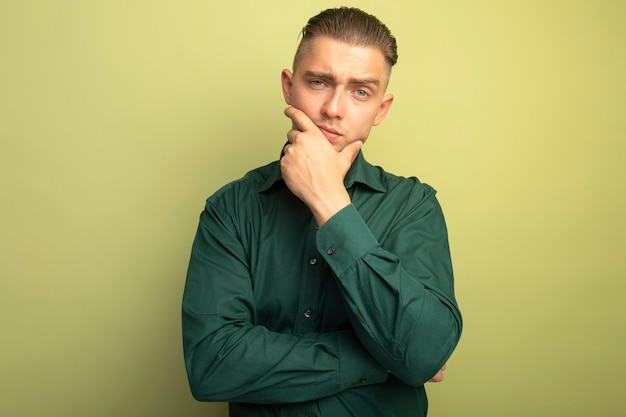 Hombre guapo joven en camisa verde con la mano en la barbilla pensando